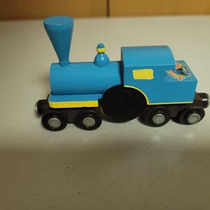 May include: A blue wooden toy train with black wheels and yellow accents. The train has a tall smokestack and a small sticker on the side. A classic toy for children.