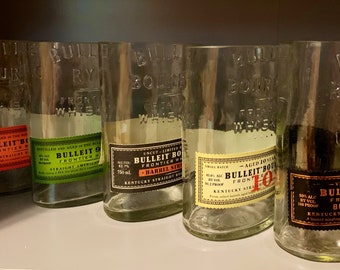 May include: Five empty glass bottles of Bulleit Bourbon Whiskey. The bottles are clear and have a textured surface. The labels are different colors and feature the Bulleit Bourbon Whiskey logo and text. The bottles are arranged in a row on a shelf.
