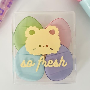 May include: A clear plastic box containing four colorful, egg-shaped makeup sponges in green, pink, blue, and purple. A yellow frog-shaped sticker with the words "so fresh" is on top. The sponges are for applying makeup.