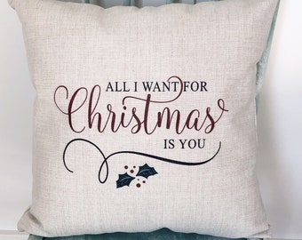 以下が含まれることがあります： 赤い筆記体で「All I want for Christmas is you」と書かれた、ベージュのクッション。下に小さなヒイラギの枝があります。クッションは薄い緑色の椅子の上に置かれています。
