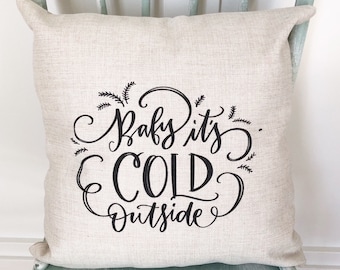 May include: A beige linen throw pillow with black text that reads "Baby it's cold outside". The pillow is sitting on a light green painted wooden chair.