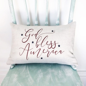 May include: A white pillow with the text "God Bless America" in red cursive lettering and blue stars. The pillow is sitting on a light green painted chair.