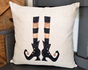 May include: A beige pillow with a black and orange witch's legs and shoes design. The pillow is on a dark gray bench.