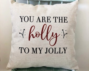 以下が含まれることがあります： 薄いベージュのクッションに、「You are the holly to my jolly」という黒い文字と赤い文字で書かれたテキストがあります。クッションは薄い青色の木製の椅子の上に置かれています。