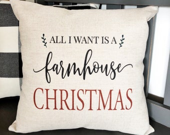 以下が含まれることがあります： 黒と赤の文字で「ALL I WANT IS A farmhouse CHRISTMAS」と書かれたベージュのクッション。クッションは、黒と白のチェック柄のクッションの後ろにある灰色のベンチの上に置かれています。