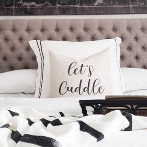 Puede incluir: Una cama blanca con una manta de rayas blancas y negras y una almohada blanca con el texto "Let's Cuddle" en negro. La cama tiene una cabecera capitoné y una colcha blanca. Hay una mesa de bandeja de metal negro en la cama con una jarra blanca y una pequeña planta.