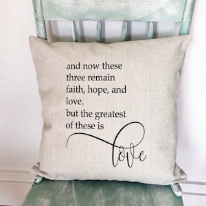 May include: A beige throw pillow with the text "and now these three remain faith, hope, and love. but the greatest of these is love" in black lettering. The pillow is sitting on a light green wooden chair.