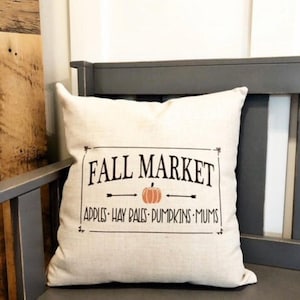 May include: A beige throw pillow with a black and white graphic design that says "Fall Market" with an arrow pointing to a pumpkin. The text below the graphic says "Apples - Hay Bales - Pumpkins - Mums".