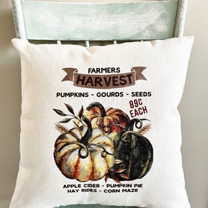 May include: A white pillow with a brown banner that says "Farmers Harvest" and a graphic of pumpkins, gourds, and seeds. The text below the graphic says "Pumpkins - Gourds - Seeds 89c Each Apple Cider - Pumpkin Pie Hay Rides - Corn Maze @Our Rustic Home Decor"