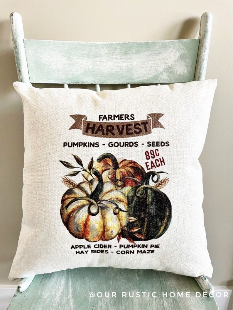 May include: A white pillow with a brown banner that says "Farmers Harvest" and a graphic of pumpkins, gourds, and seeds. The text below the graphic says "Pumpkins - Gourds - Seeds 89c Each Apple Cider - Pumpkin Pie Hay Rides - Corn Maze @Our Rustic Home Decor"