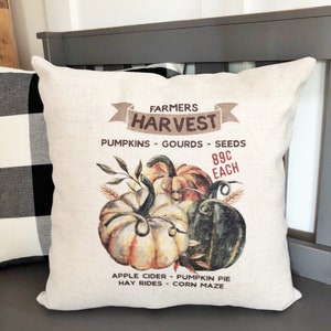 May include: A beige pillow with a brown and white illustration of pumpkins, gourds, and seeds. The text "Farmers Harvest" is in a brown banner above the pumpkins. The text "Pumpkins - Gourds - Seeds" is below the pumpkins. The text "89c Each" is in a brown rectangle to the right of the pumpkins. The text "Apple Cider - Pumpkin Pie - Hay Rides - Corn Maze" is below the pumpkins.