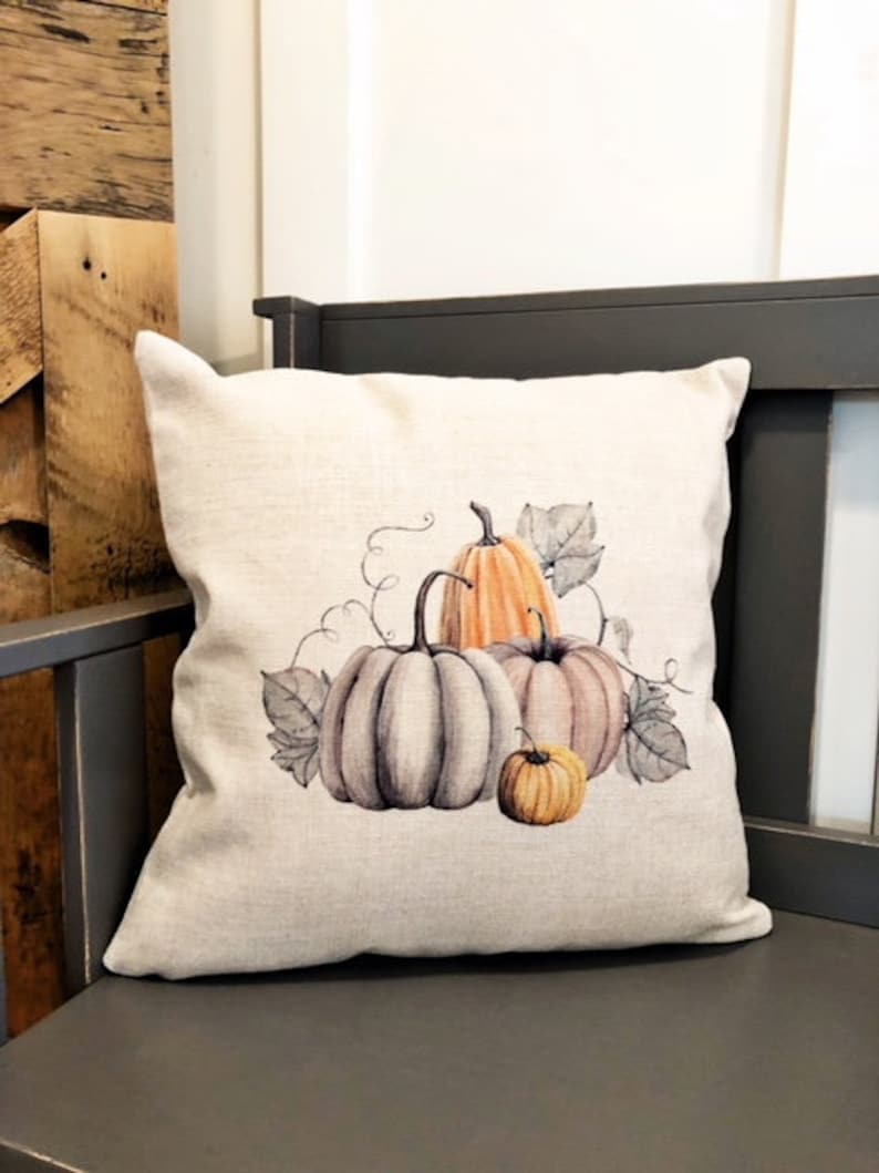 May include: A beige throw pillow with a watercolor design of three pumpkins. The pumpkins are orange, gray, and a light brown color. The pumpkins are surrounded by green leaves.