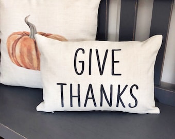 May include: Two decorative throw pillows. One pillow features a watercolor-style orange pumpkin. The other pillow is rectangular and has the words "GIVE THANKS" printed in black. The pillows are on a gray bench.