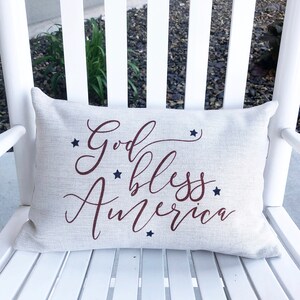May include: A white pillow with the words "God Bless America" in red cursive lettering and blue stars printed on it. The pillow is sitting on a white rocking chair.