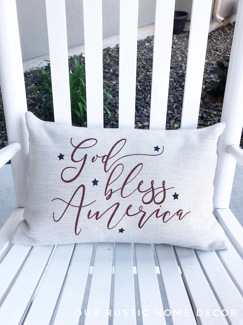 May include: A white pillow with the words "God Bless America" in red cursive lettering and blue stars printed on it. The pillow is sitting on a white rocking chair.