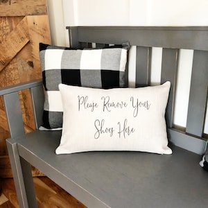 May include: A gray wooden bench with two decorative pillows. One pillow is black and white buffalo check. The other is a rectangular cream-colored pillow with the text "Please Remove Your Shoes Here" in black script.