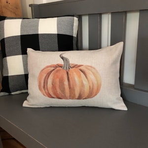 K&ouml;nnte beinhalten: Eine graue Holzbank mit zwei Kissen. Ein Kissen ist schwarz-wei&szlig; kariert und das andere ist ein hellbraunes Leinenkissen mit einer Aquarellmalerei eines orangefarbenen K&uuml;rbisses.