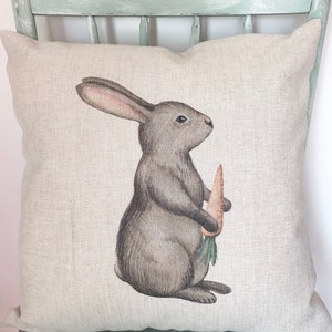 May include: A square decorative pillow with a printed illustration of a gray bunny holding a carrot. The bunny has long ears and is facing to the right. The pillow has a neutral linen-like texture.