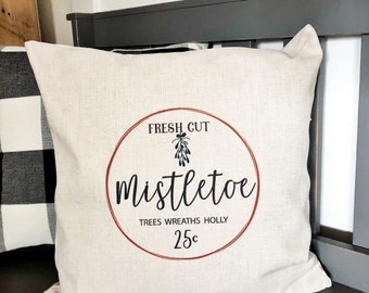 Puede incluir: Una almohada beige con un círculo rojo y el texto "FRESH CUT MISTLETOE TREES WREATHS HOLLY 25¢" impreso en ella. La almohada está sentada en un banco gris oscuro.