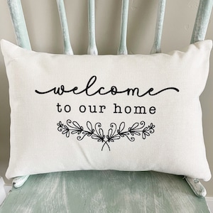 May include: White cushion with black text that reads "welcome to our home" with a decorative floral design underneath.