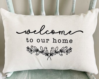Könnte beinhalten: Ein weißes Kissen mit schwarzem Text, der "Willkommen in unserem Zuhause" lautet, mit einem dekorativen floralen Design darunter.