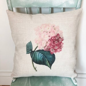 May include: A beige linen pillow with a pink and red hydrangea flower print. The pillow is sitting on a light green chair.