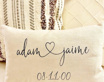 May include: A rectangular beige pillow with the names "adam & jaime" in black script, connected by a heart symbol. Below the names is the date "08.11.00". The pillow is part of a home decor collection.