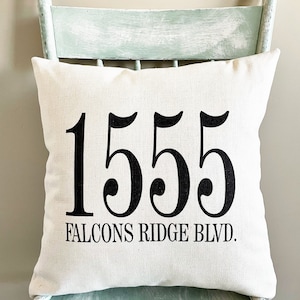 May include: Cream-colored square pillow with the address "1555 FALCONS RIDGE BLVD." printed in bold black letters. The pillow is placed on a distressed, light blue wooden chair, creating a rustic aesthetic. The pillow is a home decor item.