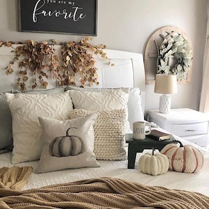 May include: A white bed with a brown knitted blanket, white pillows, and a gray pumpkin pillow. The headboard has a sign that says "dear autumn, you are my favorite". There is a wreath on the wall behind the bed.