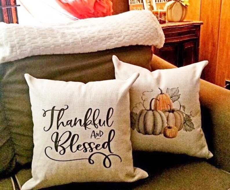 May include: Two decorative throw pillows with a fall theme. One pillow is white with black text that reads "Thankful and Blessed". The other pillow is white with a watercolor painting of pumpkins and vines.