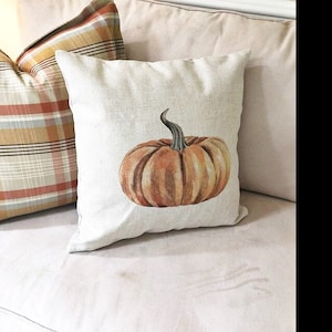 K&ouml;nnte beinhalten: Ein beiger Kissenbezug mit einem braunen und orangefarbenen K&uuml;rbisdesign. Das Kissen liegt auf einem wei&szlig;en Sofa.