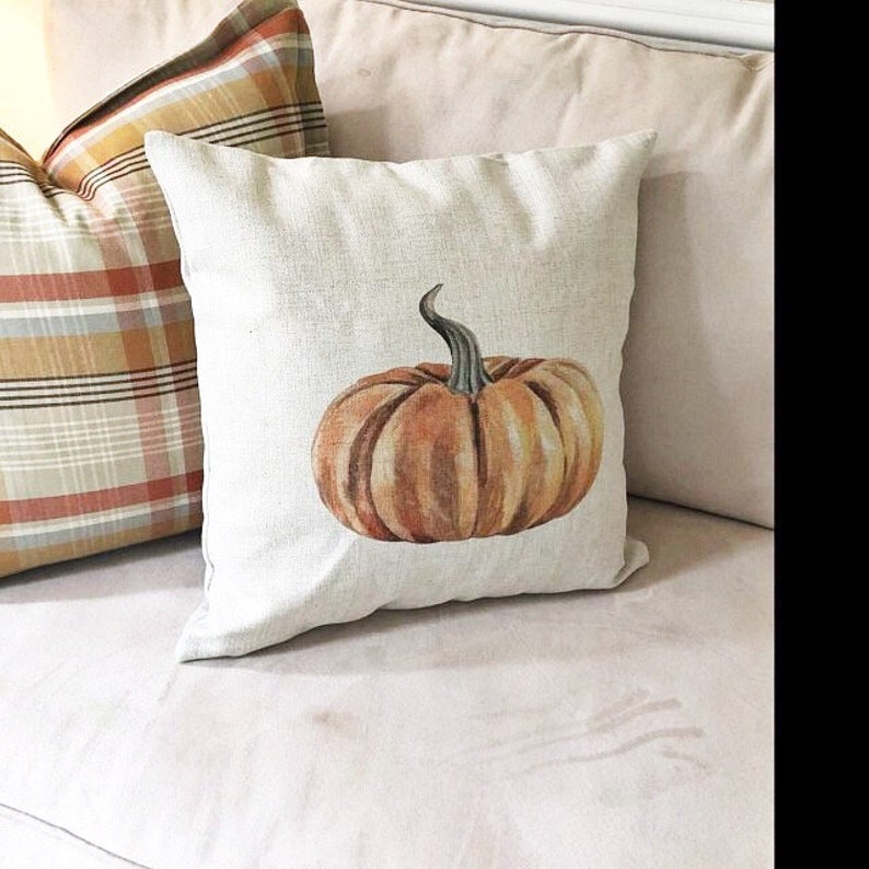 K&ouml;nnte beinhalten: Ein beiger Kissenbezug mit einem braunen und orangefarbenen K&uuml;rbisdesign. Das Kissen liegt auf einem wei&szlig;en Sofa.