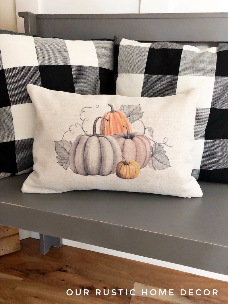 May include: A decorative throw pillow with a watercolor design of pumpkins and leaves. The pillow is beige and has a subtle texture. The pumpkins are orange, gray, and brown. The leaves are green and brown.