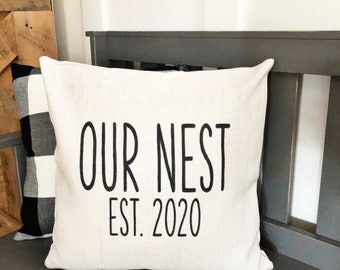 May include: A white throw pillow with black text that reads "OUR NEST EST. 2020". The pillow is sitting on a gray bench.