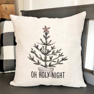Puede incluir: Una almohada blanca con una ilustración de acuarela en blanco y negro de un árbol de Navidad con una estrella rosa en la parte superior. El texto "OH HOLY NIGHT" está impreso debajo del árbol.