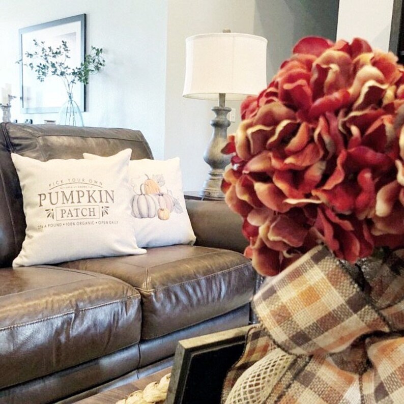 May include: A brown leather couch with two throw pillows. One pillow has a white background with a watercolor illustration of pumpkins and the text "Pick Your Own Pumpkin Patch". The other pillow has a white background with a brown and white plaid pattern. A bouquet of red flowers is in the foreground.