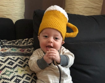 May include: A crocheted hat shaped like a beer mug with a yellow body, a white textured rim representing foam, and a yellow handle on the side. The hat is worn by a baby dressed in a beige and white striped long-sleeve onesie with gray cuffs.