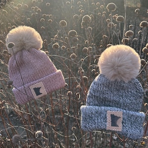 May include: Two knitted beanies with faux fur pom-poms. One is light pink, the other is gray. Each beanie has a leather patch with a cutout of the state of Minnesota.