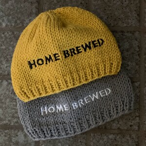 May include: Two knit beanies, one mustard yellow and one gray, each featuring the text "HOME BREWED" embroidered on the front. The yellow beanie has black lettering, while the gray beanie has white lettering, both in a chunky knit style.
