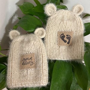 May include: Two off-white knitted beanies with small rounded ears on top. Each hat features a square tan patch stitched to the front. One patch displays the text "special delivery" in black script, while the other shows two black baby footprints.