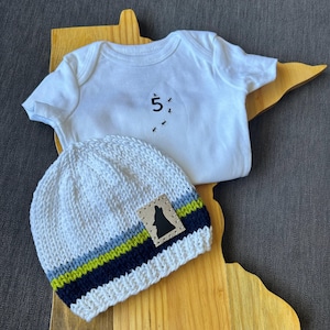 Könnte beinhalten: Ein weißer Baby-Body mit der Zahl "5" und kleinen schwarzen Insekten-Grafiken, kombiniert mit einer weißen Strickmütze. Die Mütze hat einen Lederaufnäher mit einer Wolfssilhouette und blauen, grünen und marinefarbenen Streifen.