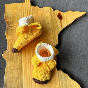 May include: Two handmade, knitted banana-shaped cozies, one upright and one open, with white and brown accents. The cozies are on a wooden cutting board shaped like the state of Minnesota. The background is a gray surface.