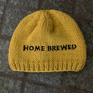 May include: A bright yellow knit beanie featuring the text HOME BREWED embroidered in black thread across the front. The hat is constructed with a thick, chunky yarn texture and a ribbed hem at the base, showcasing a simple, rounded silhouette.