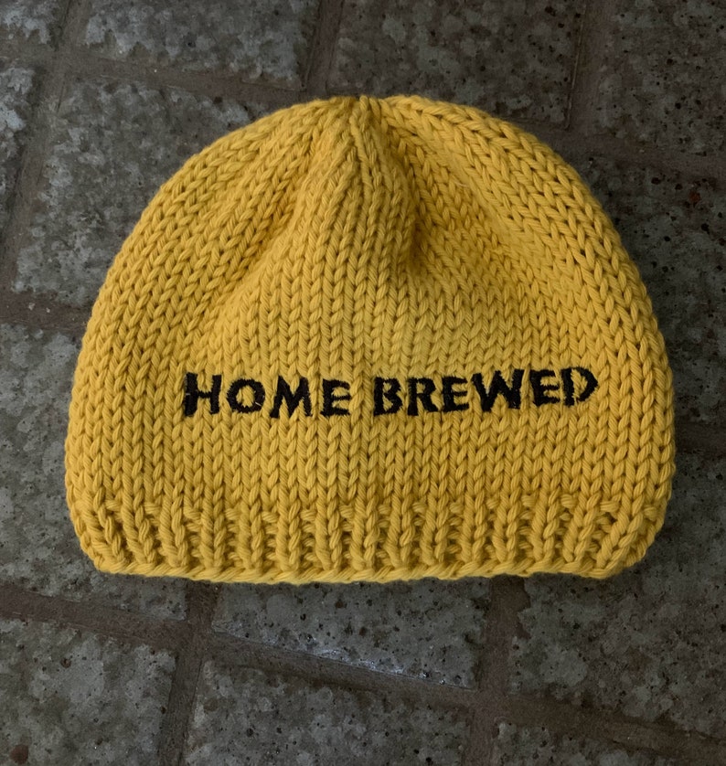 May include: A bright yellow knit beanie featuring the text HOME BREWED embroidered in black thread across the front. The hat is constructed with a thick, chunky yarn texture and a ribbed hem at the base, showcasing a simple, rounded silhouette.