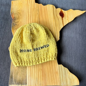 May include: A mustard yellow knit beanie with a ribbed brim and the text HOME BREWED embroidered in black thread on the front, resting on a light-colored wooden board shaped like the outline of the state of Minnesota.