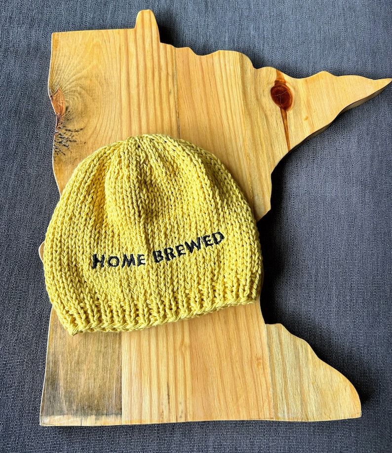 May include: A mustard yellow knit beanie with a ribbed brim and the text HOME BREWED embroidered in black thread on the front, resting on a light-colored wooden board shaped like the outline of the state of Minnesota.