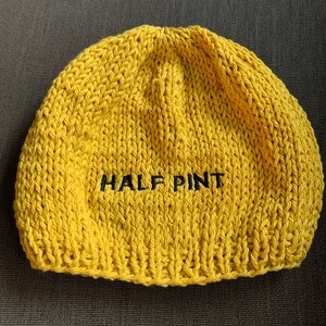 May include: A bright yellow knit beanie hat featuring the text HALF PINT embroidered in black thread across the center. The hat is constructed with a chunky yarn texture and a standard rounded crown shape, showcasing a simple design.