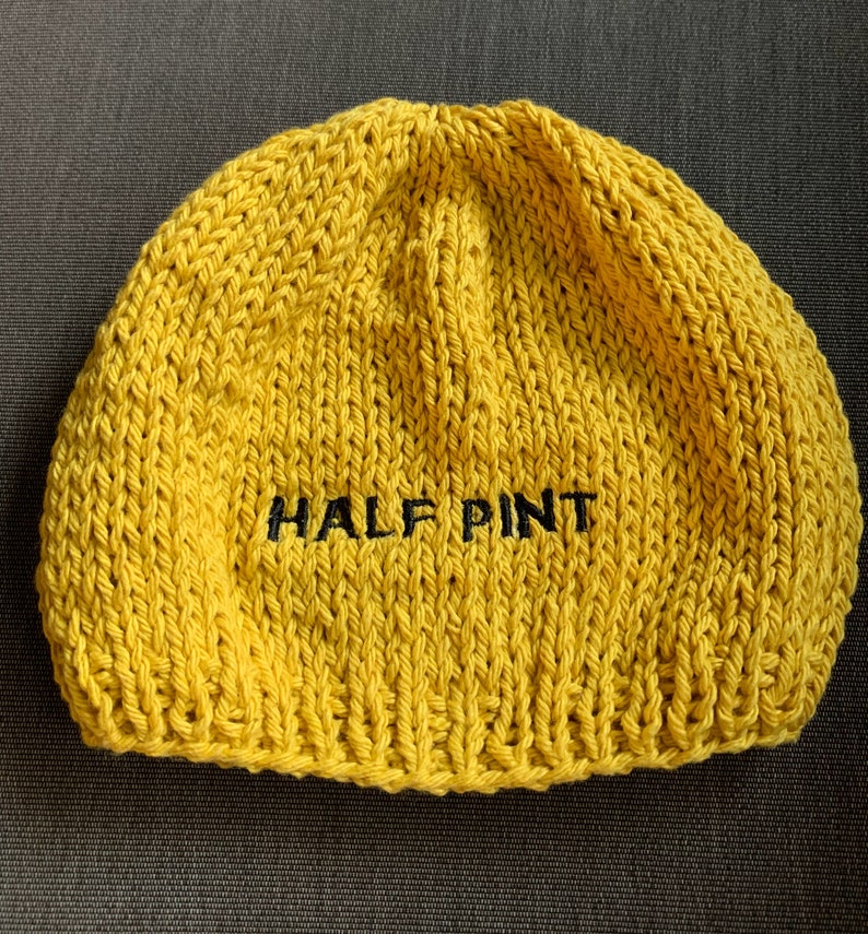 May include: A bright yellow knit beanie hat featuring the text HALF PINT embroidered in black thread across the center. The hat is constructed with a chunky yarn texture and a standard rounded crown shape, showcasing a simple design.