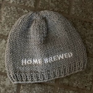 May include: A light gray knit beanie featuring the text HOME BREWED embroidered in white thread across the front. The hat is constructed with a thick, textured yarn in a standard stockinette stitch pattern with a ribbed edge at the bottom.