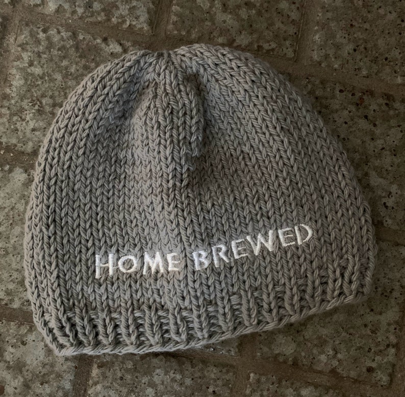 May include: A light gray knit beanie featuring the text HOME BREWED embroidered in white thread across the front. The hat is constructed with a thick, textured yarn in a standard stockinette stitch pattern with a ribbed edge at the bottom.